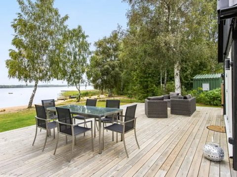 Große Holzterrasse mit Esstisch, Stühlen und Lounge-Bereich am Wasser mit Blick auf den See, umgeben von Bäumen.