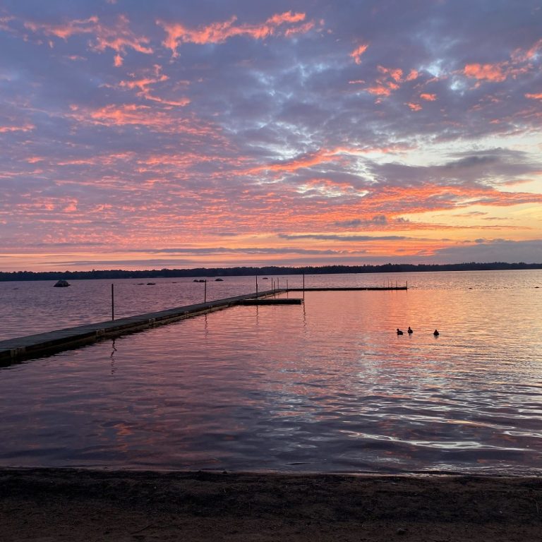 Sonnenuntergang über dem Wasser mit lila schimmernden Wolken und einem Steg im Wasser.