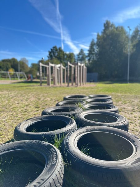 Reihen von alten Reifen auf einer Spielplatzwiese mit Klettergeräten im Hintergrund.