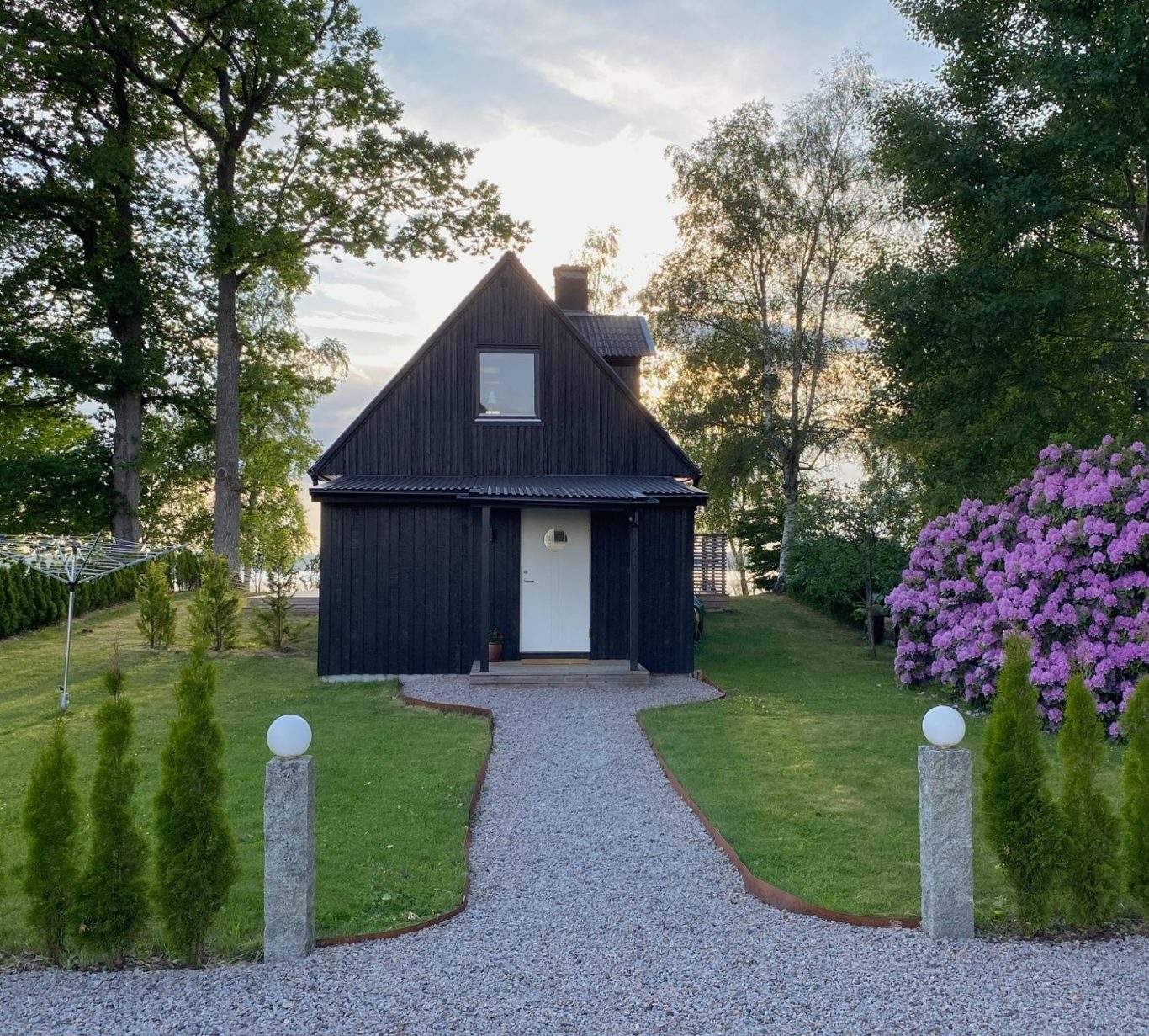 Schwarzes Holzhaus mit Garten und lila Rhododendron, umgeben von Bäumen und einem Kiesweg, im Hintergrund der See.